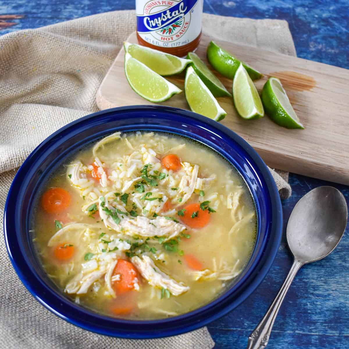 The chicken and rice soup served in a blue bowl and set on a blue table with a beige linen and lime wedges in the background.