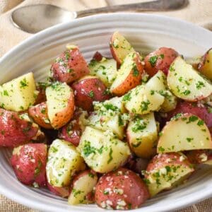 The finished garlic parsley potatoes served in a white bowl on a beige linen with a serving spoon in the background.
