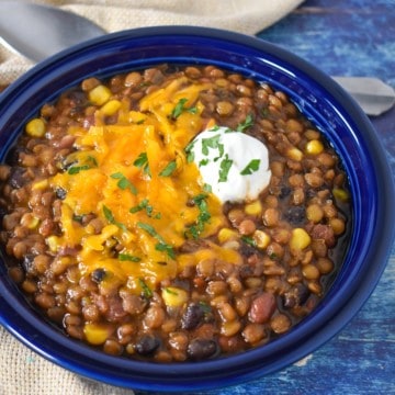The chili served in a blue bowl and garnished with melted cheddar cheese and a dollop of sour cream displayed on a blue table with a beige linen and a spoon.