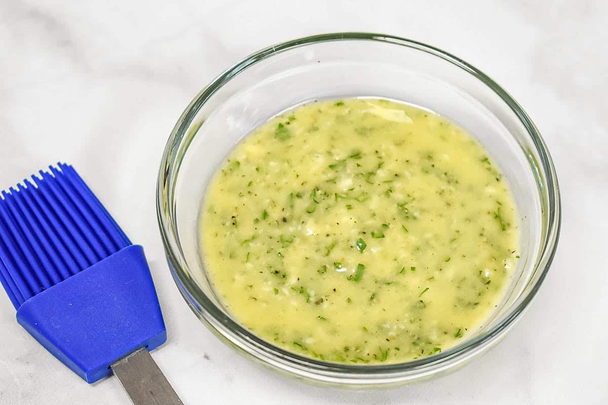 Garlic butter mixture with olive oil, fresh garlic, parsley, salt, and black pepper in a glass bowl, with a blue pastry brush on the side.
