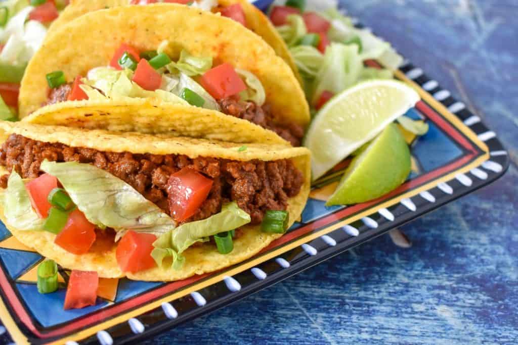 Two tacos in a hard shell topped with lettuce and tomatoes displayed on a colorful plate on a blue table.
