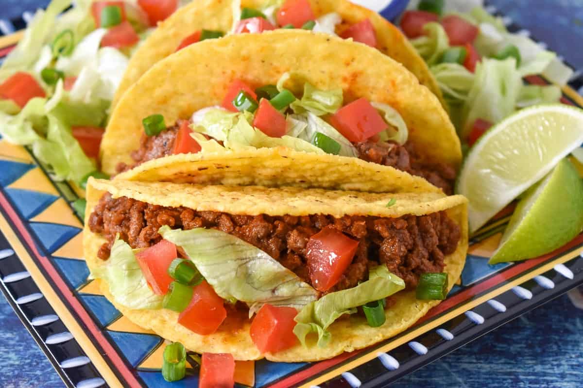 The ground beef in hard shell tacos topped with diced tomatoes, lettuce and green onions served on a colorful plate and set on a blue table.