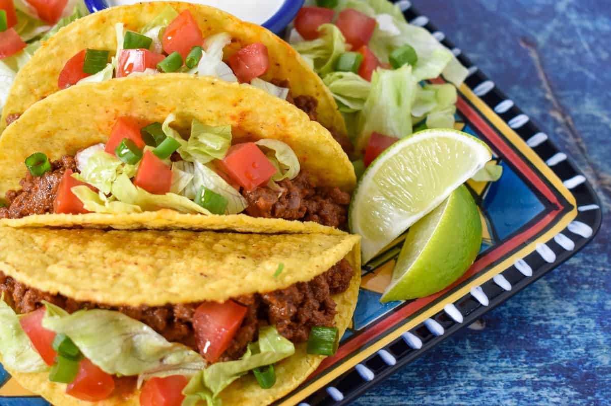 Three tacos topped with lettuce, tomato and green onions displayed on a colorful plate with two lime wedges to the right and set on a blue table.