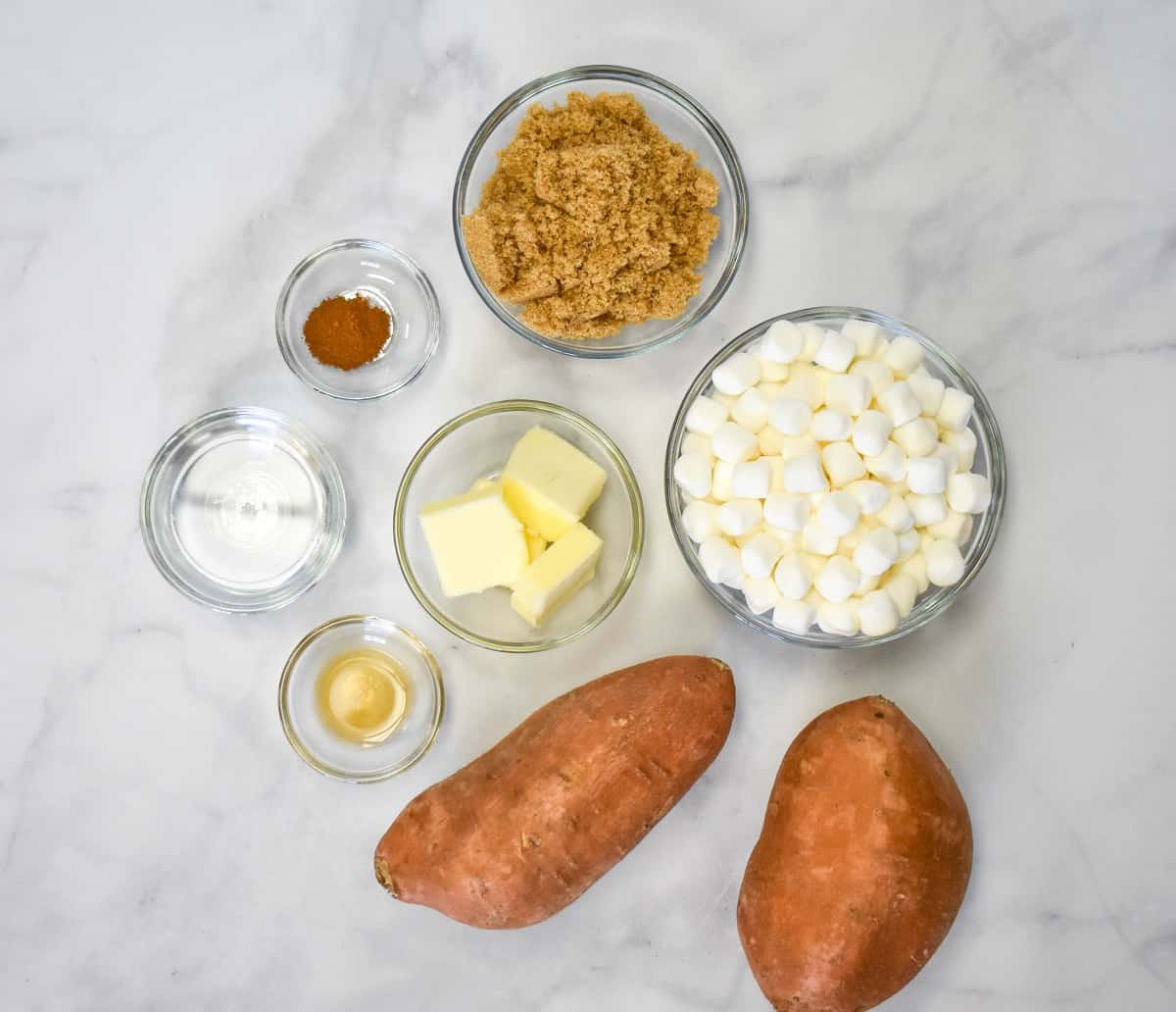 Two sweet potatoes and the rest of the ingredients for the recipe in clear glass bowls all arranged on a white table.