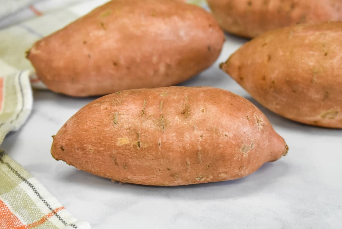 Four sweet potatoes arranged on a white table with a light colored kitchen towel on the left side.