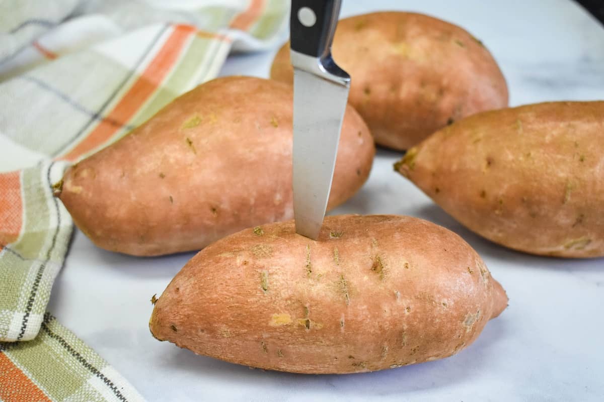 Four sweet potatoes on a white table the one in the forefront is being pricked with a paring knife.