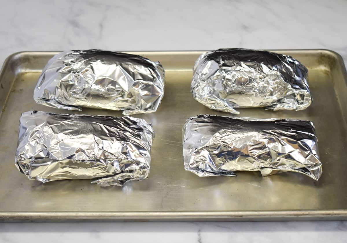 Four sweet potatoes wrapped in foil and arranged on a baking sheet.