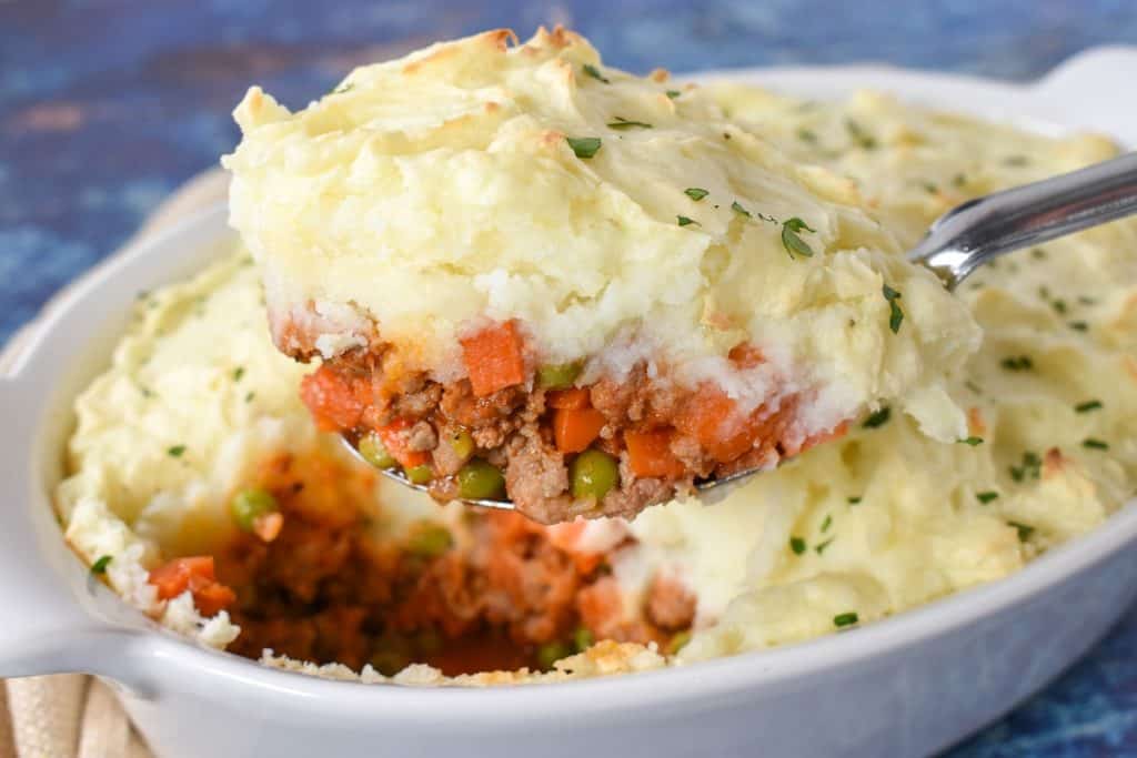 A serving spoon lifting a serving of shepherd's pie out of a white casserole dish.