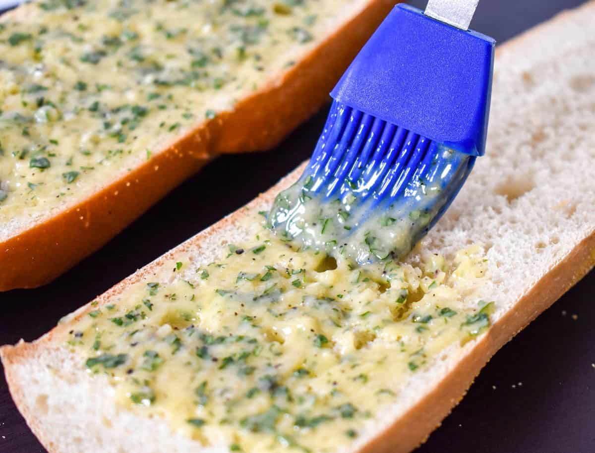 Garlic butter being spread over sliced French bread with a blue pastry brush.