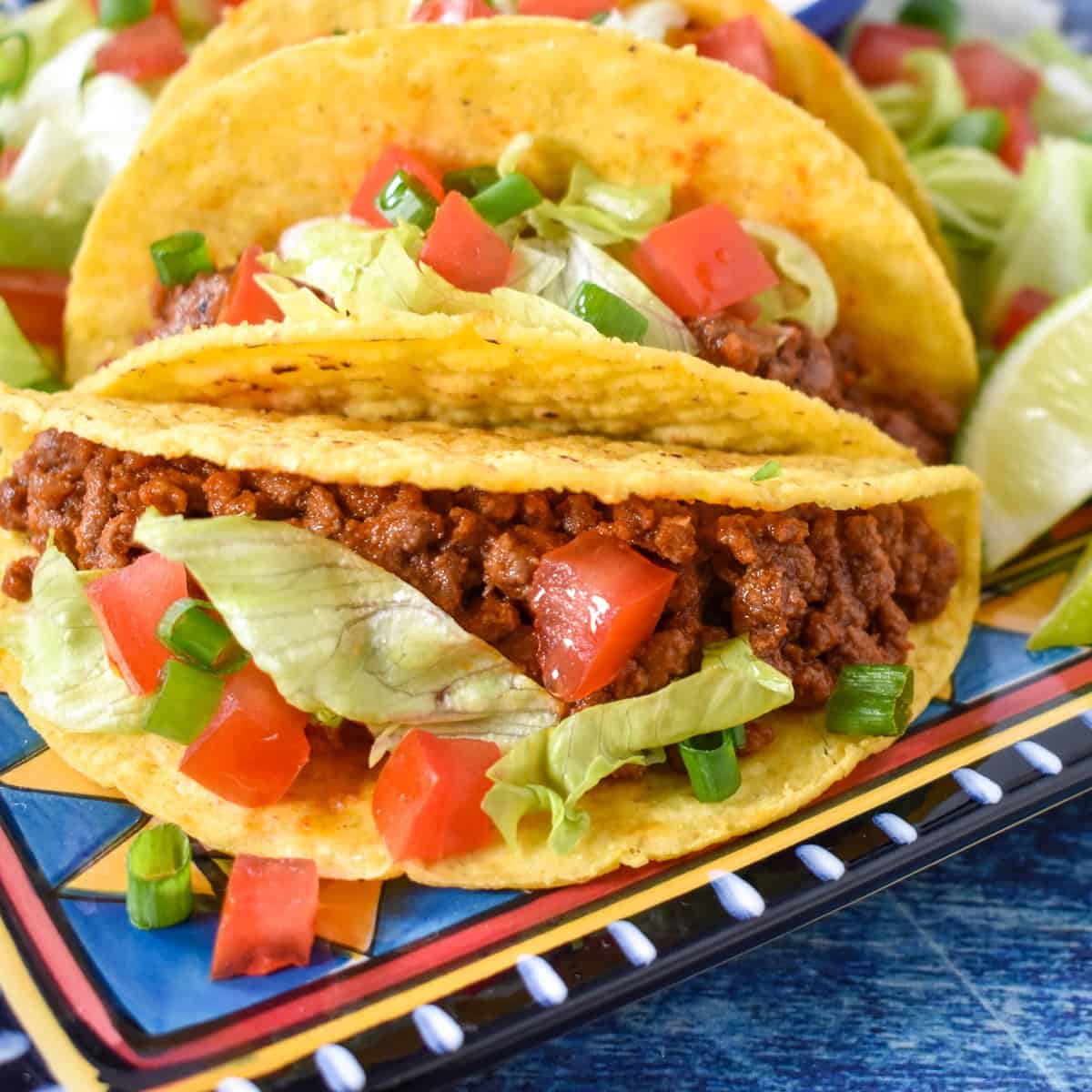 Two tacos in a hard shell topped with lettuce and tomatoes displayed on a colorful plate on a blue table.