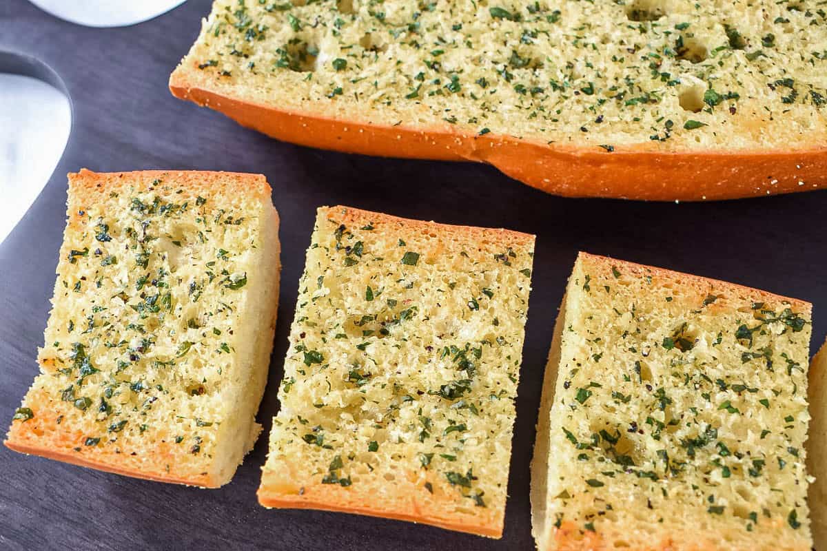 Slices of homemade garlic bread with golden toasted edges and parsley on a dark cutting board.