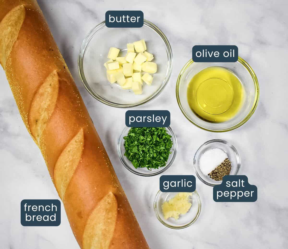 The ingredients for the garlic bread, prepped and arranged in white bowls set on a white table, with each labeled.
