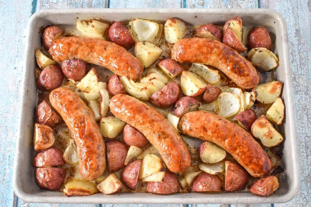 An image of five Italian sausages cooked on a sheet pan with quartered red potatoes and onions.