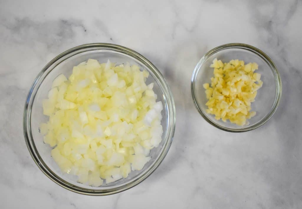 Diced onions and minced garlic displayed in glass bowls on a white table.