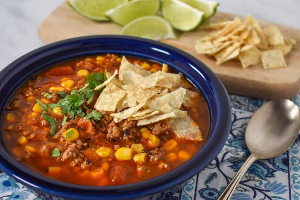 The soup served in a blue plate and displayed on a blue and white linen with lime wedges and extra tortilla chips in the background.