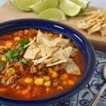 The soup served in a blue plate and displayed on a blue and white linen with lime wedges and extra tortilla chips in the background.