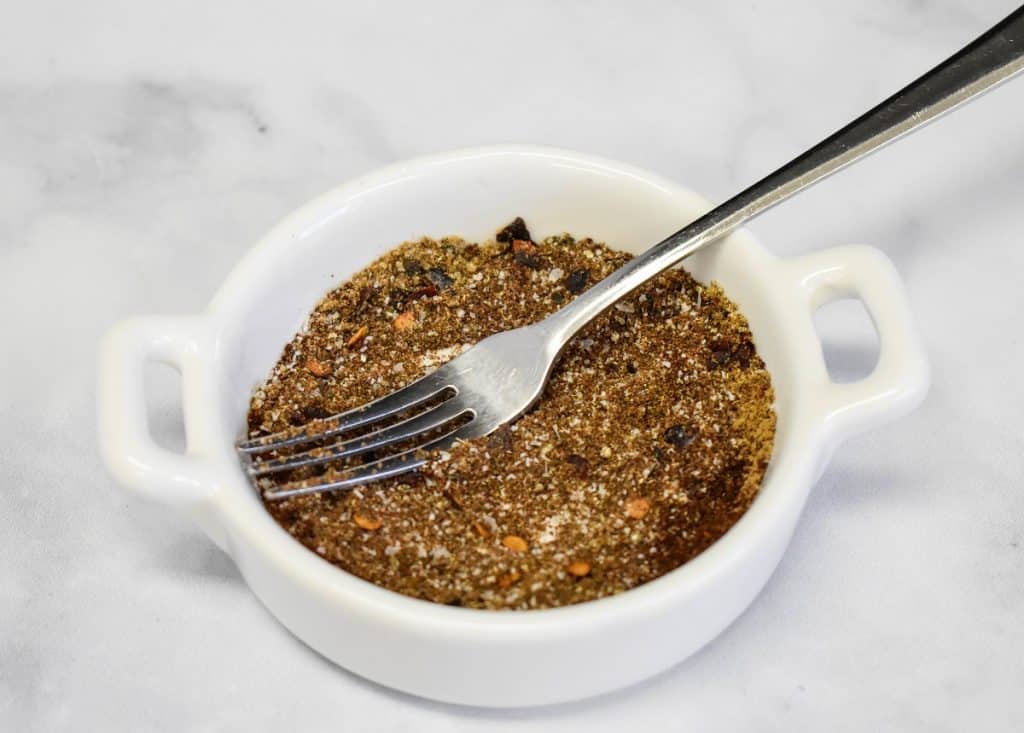 The seasoning mix in a small white bowl and a small cocktail fork displayed on a white table.