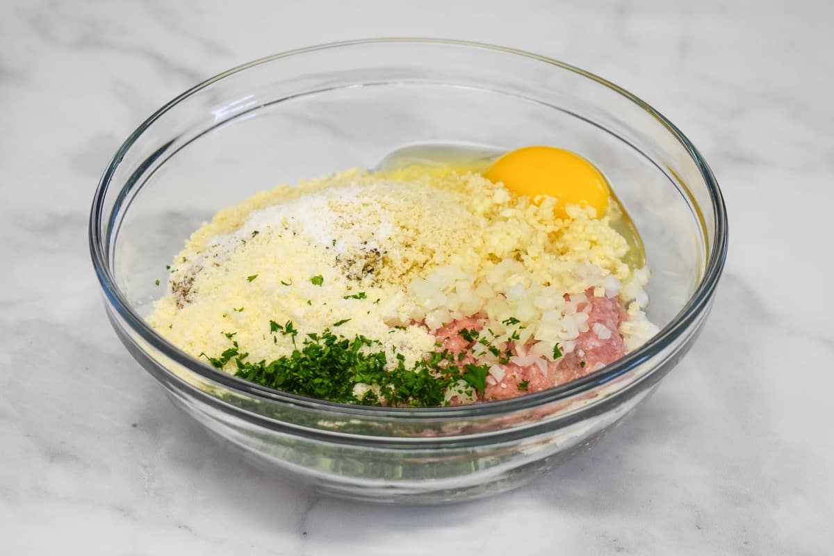 The ingredients for the meatballs added to a large glass bowl set on a white table.