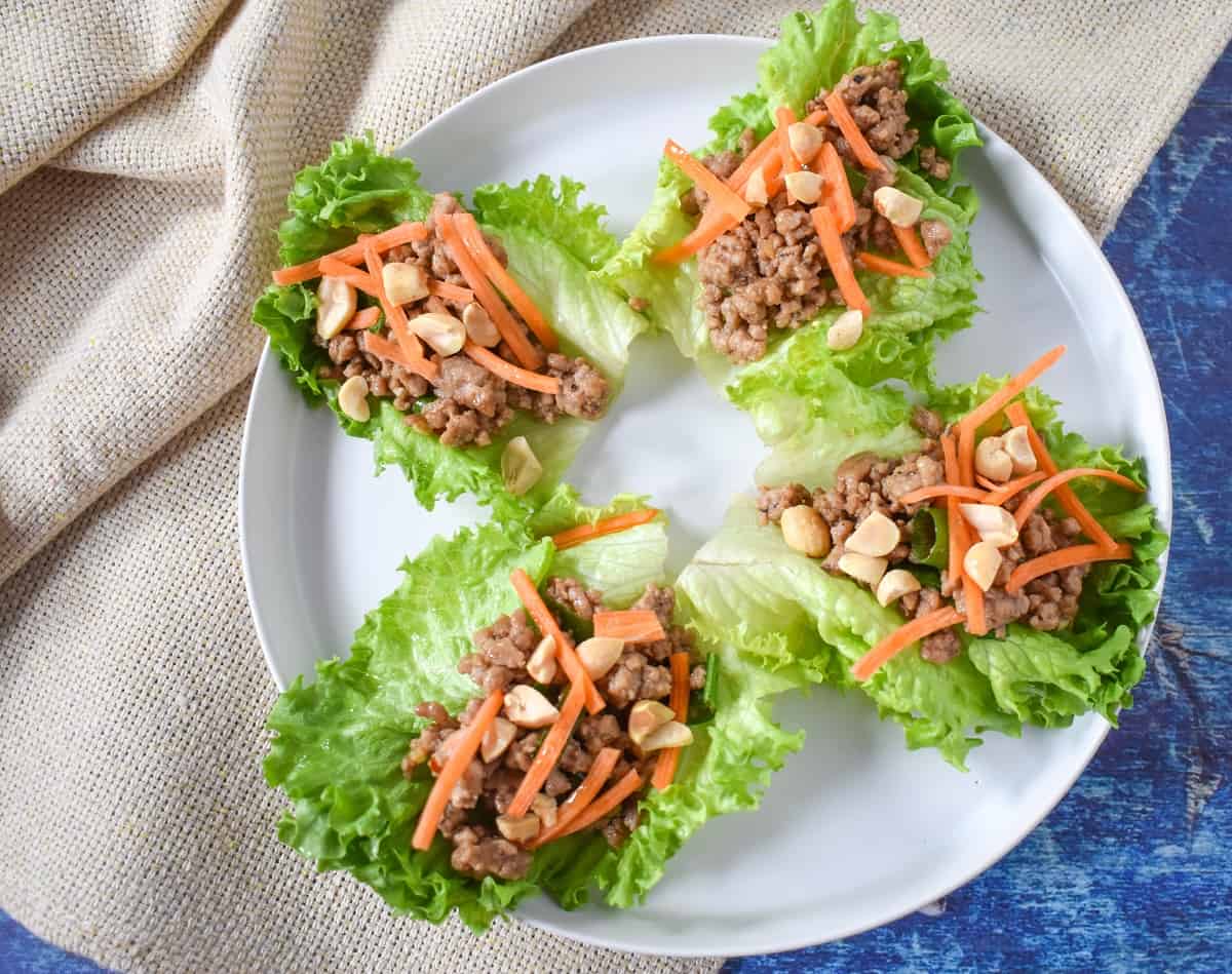 Four prepared lettuce cups served on a white plate and set on a beige linen on a blue table.