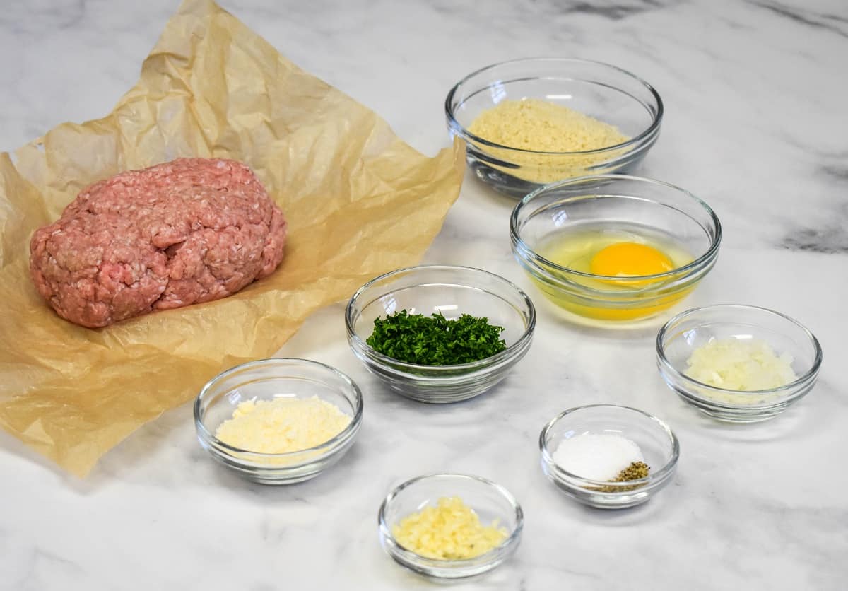 The prepped ingredients for the meatballs arranged in small glass bowls on a white table.