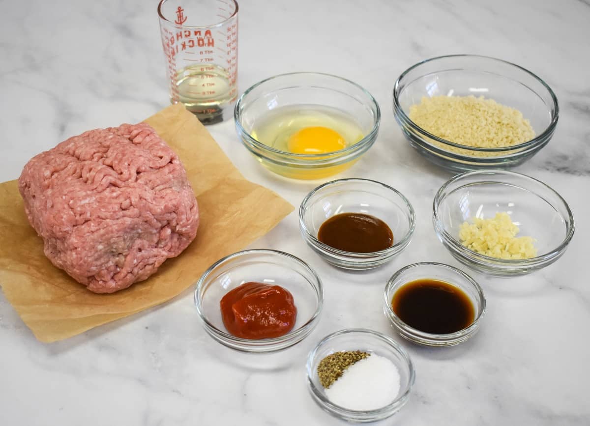 The ingredients for the turkey Salisbury steak patties arranged in clear bowls, the turkey on parchment paper, on a white table.