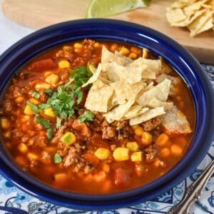 The soup served in a blue plate and displayed on a blue and white linen with lime wedges and extra tortilla chips in the background.