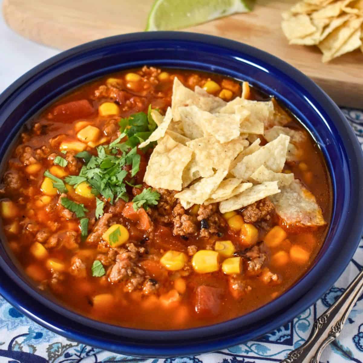 The soup served in a blue plate and displayed on a blue and white linen with lime wedges and extra tortilla chips in the background.