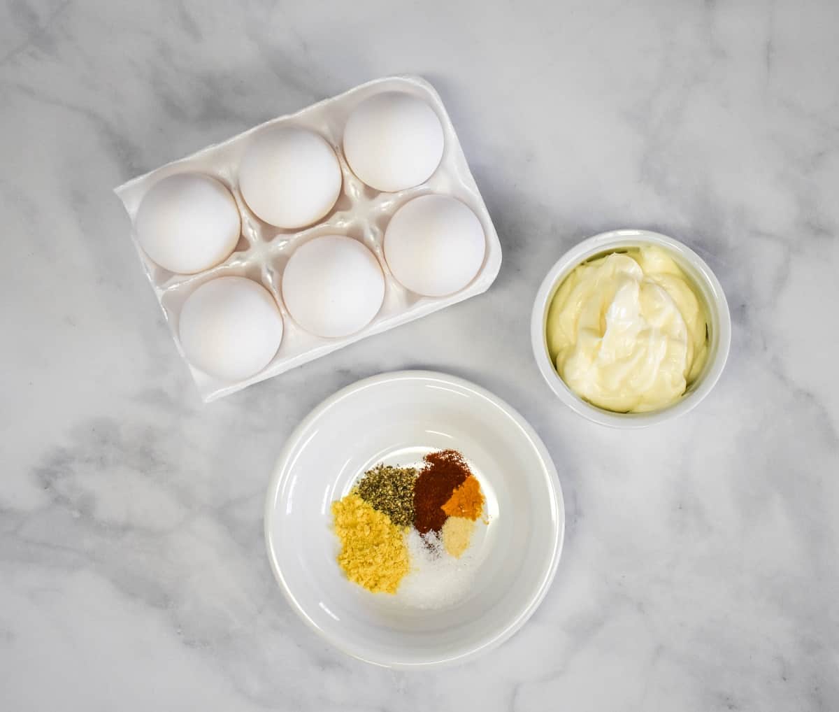 The ingredients for the egg salad arranged on a white table. The spices are displayed on a small white plate.