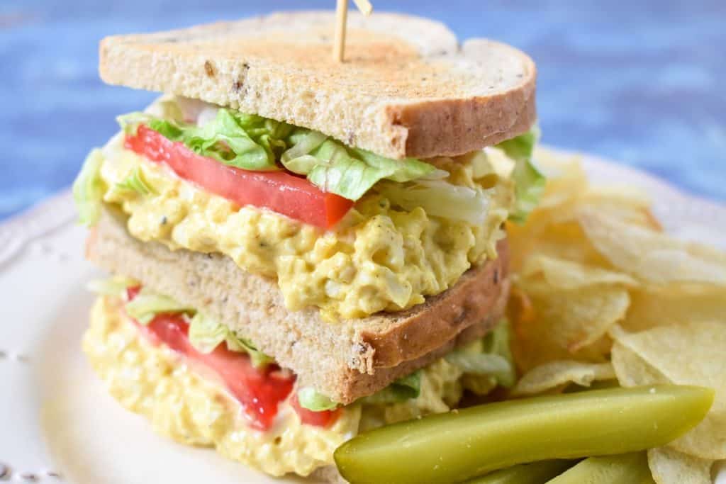 A sandwich made with the egg salad with lettuce and tomatoes and rye bread. The sandwich is displayed on a white plate with chips and pickles on the side.