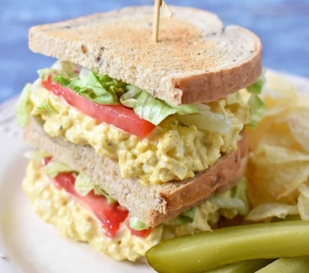 A stacked egg salad sandwich with lettuce and tomatoes on a white plate with chips and pickle spears on the side.