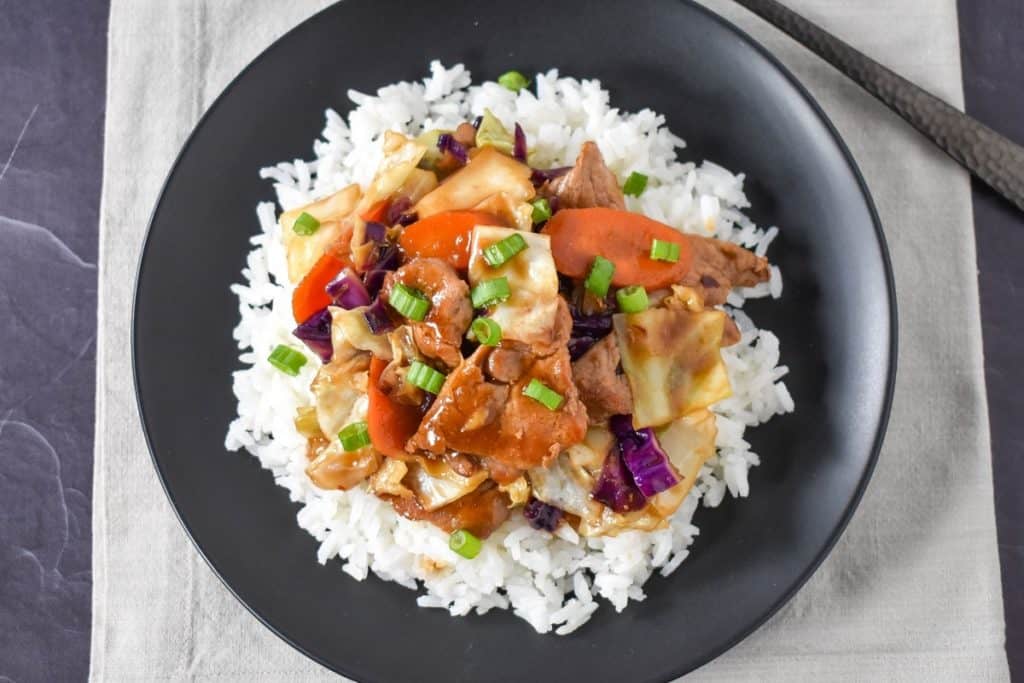 Pork stir fry served on a bed of white rice on a black plate and displayed on a beige linen.