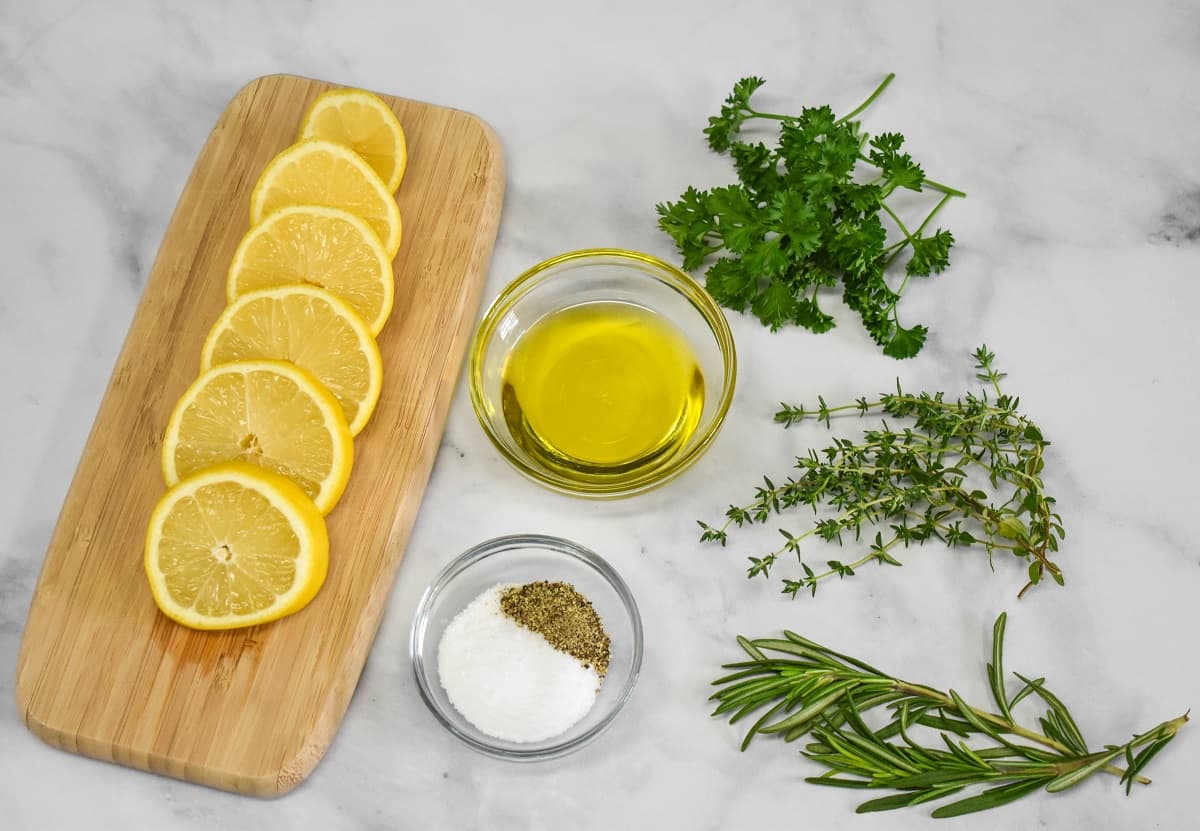 Lemon slices, herbs, olive oil, salt and pepper arranged on a white table. The lemons are displayed on a small wood cutting board.