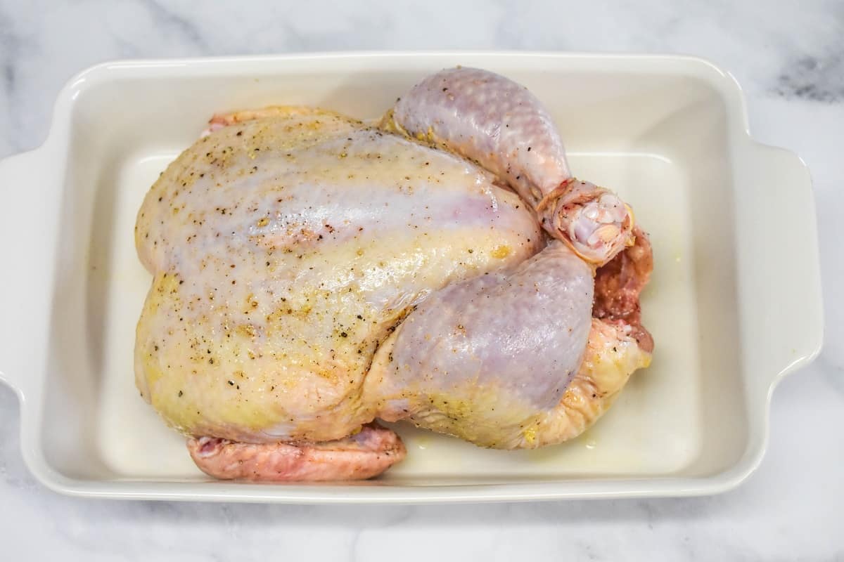 A raw, whole chicken in a white casserole dish set on a white table.