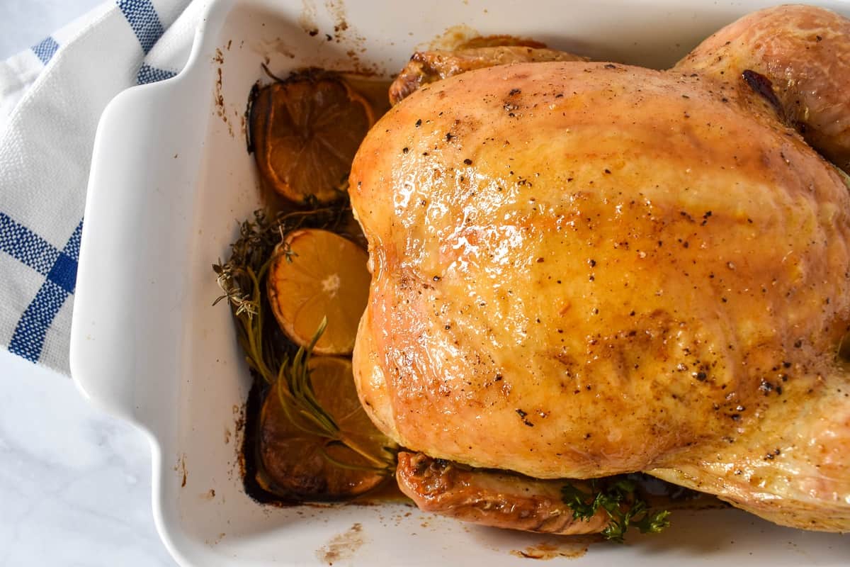 A close up of the simple roasted chicken with showing mostly the chicken breast in a white casserole dish with the pan juices, herbs and lemon slices.