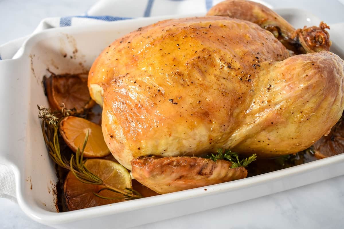 An image of the whole roasted chicken in a white casserole dish in the pan juices with the roasted lemon slices and herbs arranged around it.