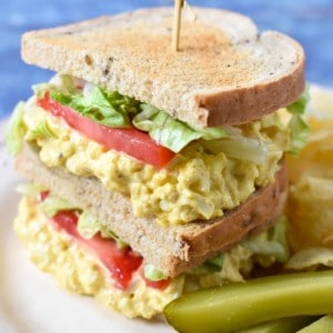 An image of a sandwich made with the egg salad with lettuce and tomatoes and rye bread. The sandwich is displayed on a white plate with chips and pickles on the side.