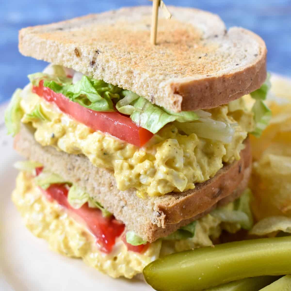 An image of a sandwich made with the egg salad with lettuce and tomatoes and rye bread. The sandwich is displayed on a white plate with chips and pickles on the side.