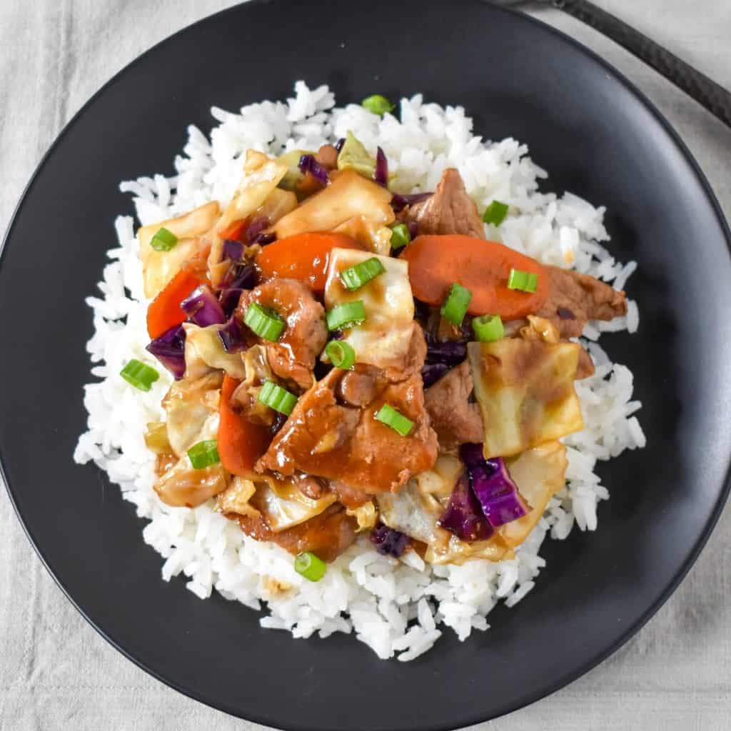 Pork stir fry served on a bed of white rice on a black plate and displayed on a beige linen.