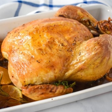 The whole roasted chicken in a white casserole dish and the pan juices and roasted lemon slices and herbs.