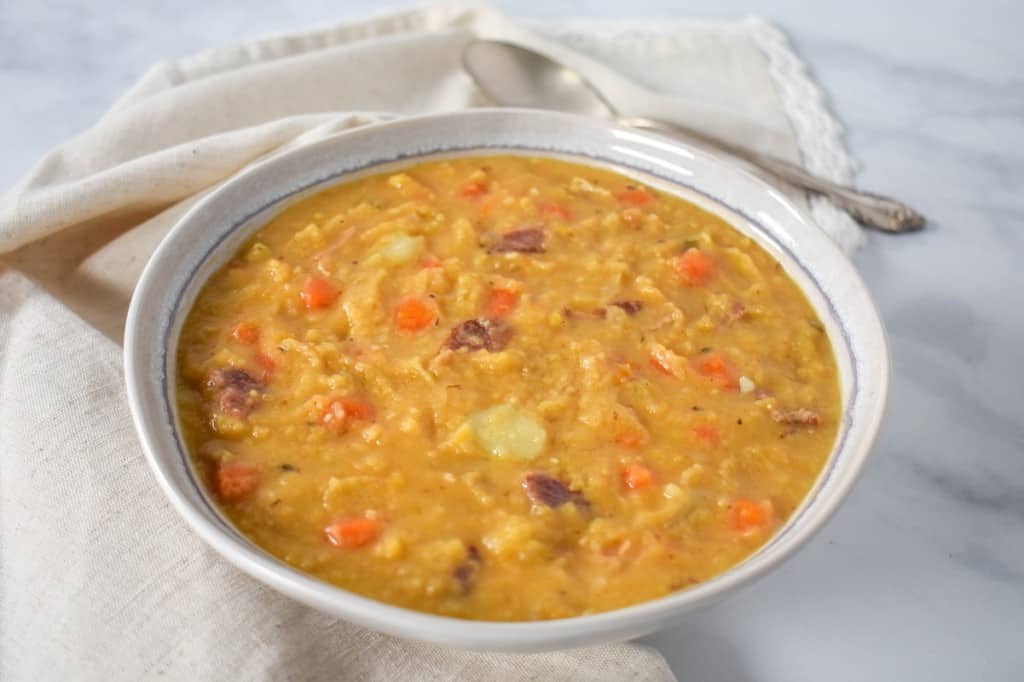 A close up of the yellow split pea soup served in a white bowl with a light beige linen with a spoon to the top right side, set on a white table..