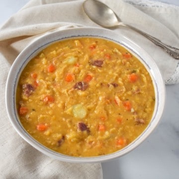 The yellow split pea soup served in a white plate with a light beige linen with a spoon to the top right side. The plate, linen and spoon are set on a white table.