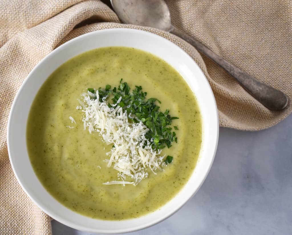 Zucchini soup garnished with chopped parsley and grated parmesan cheese. The soup is served in a white bowl with a beige linen.