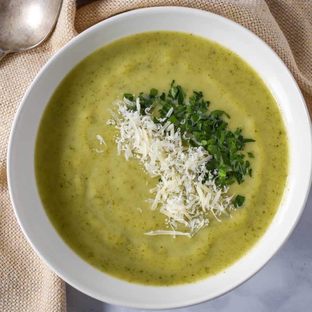 Zucchini soup garnished with chopped parsley and grated parmesan cheese. The soup is served in a white bowl with a beige linen.