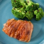 The bacon wrapped chicken thighs served on a aqua colored plate with a side of steamed broccoli garnished with a little crushed red pepper.