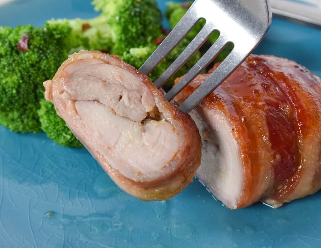 A close up of a sliced piece of chicken held up by a fork. In the background is the remaining chicken and steamed broccoli served on an aqua colored plate.