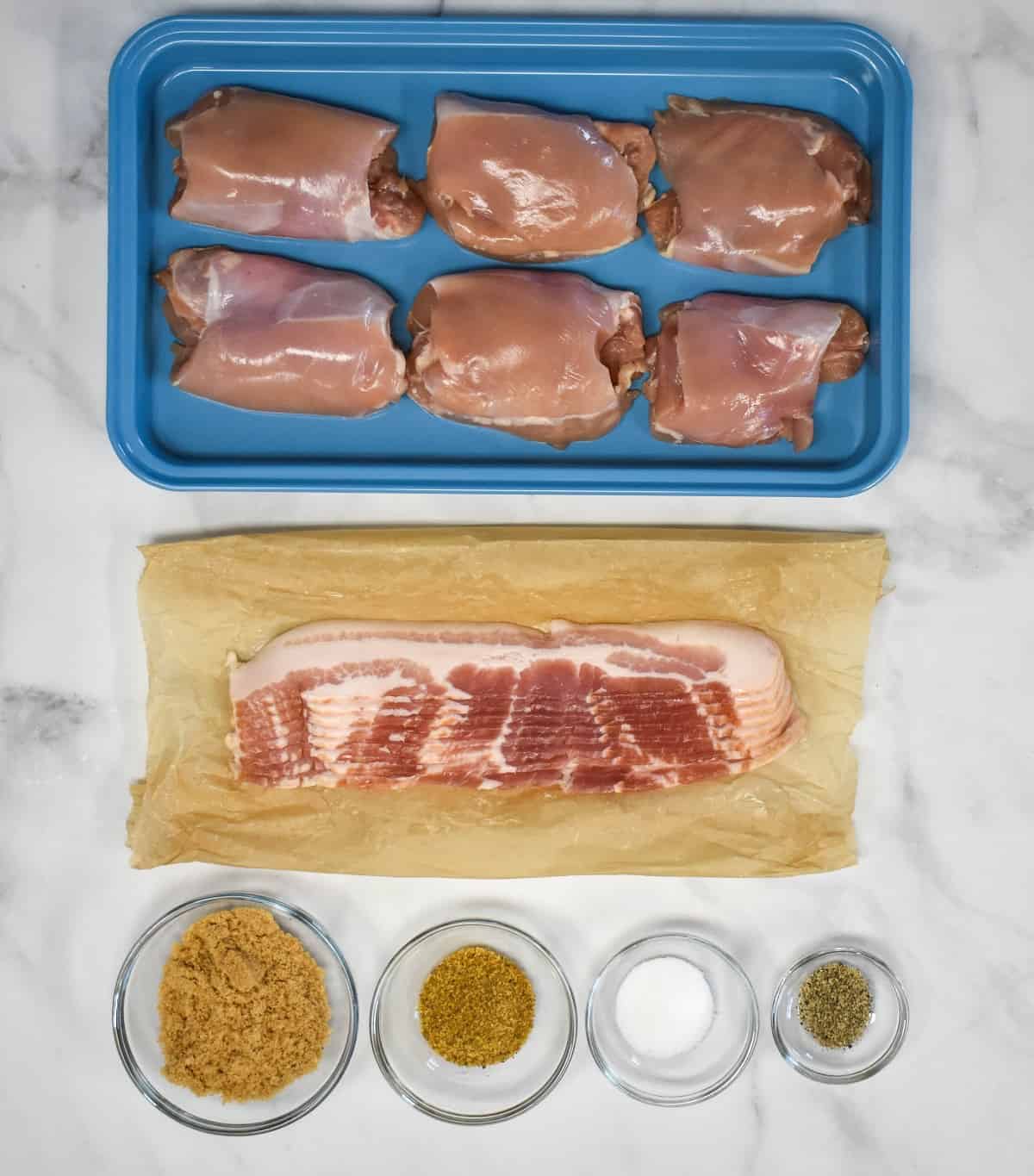 The ingredients for the dish arranged on a white table. The chicken is in a blue pan, the bacon on parchment paper and the seasoning in small glass bowls.