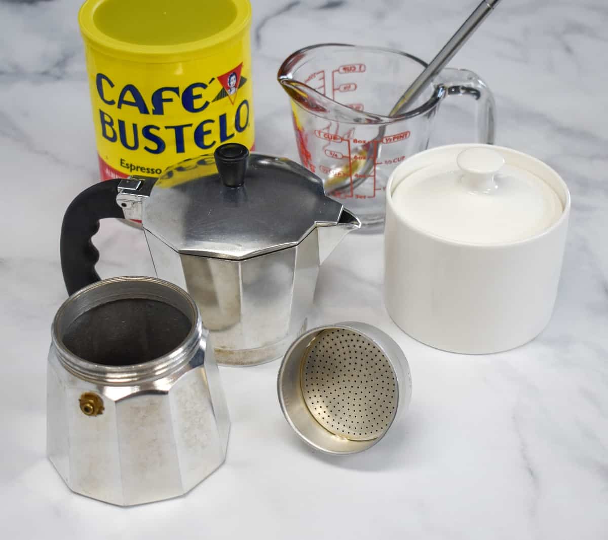 The ingredients and appliance used to make Cuban coffee arranged on a white table.