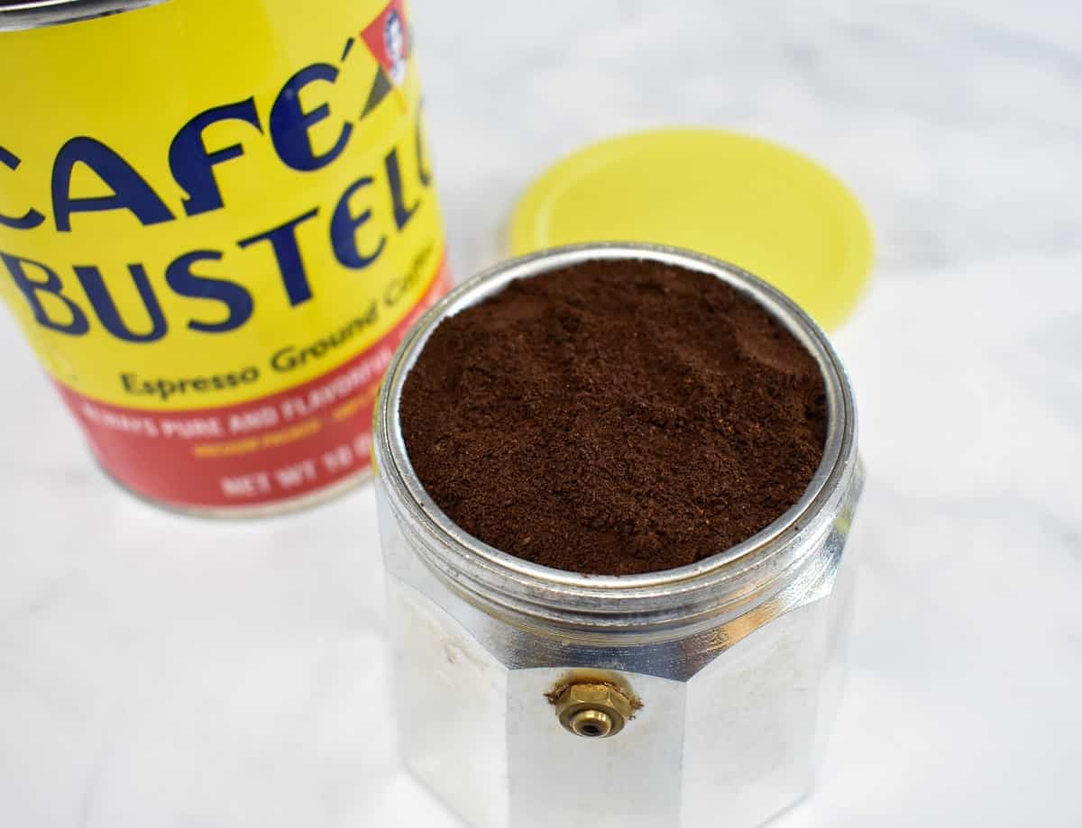 The coffee pot base with the filter inserted filled with the espresso grounds. There is a yellow tin can of coffee in the background.