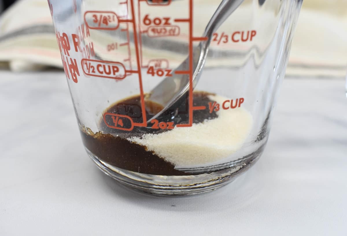Sugar and a little bit of coffee in a glass measuring cup. The cup has a spoon in it and is set on a white table.