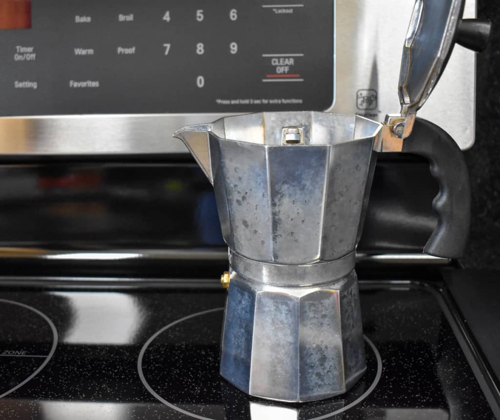 A moka coffee pot with the lid open set on a burner on a black stove.
